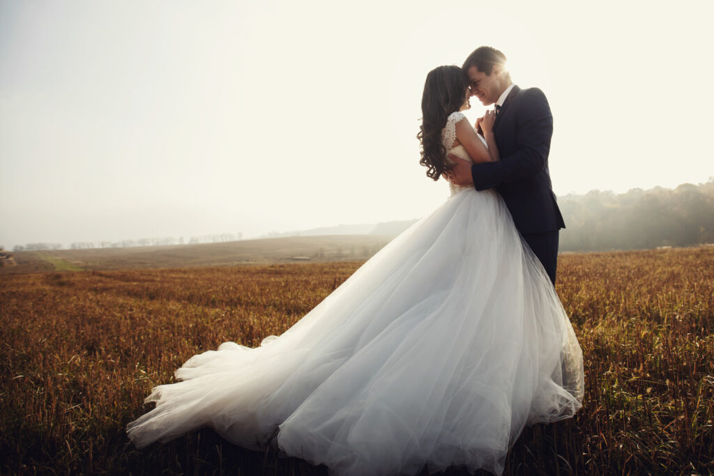 Romantic fairytale newlywed couple hug & kiss in field at sunset.