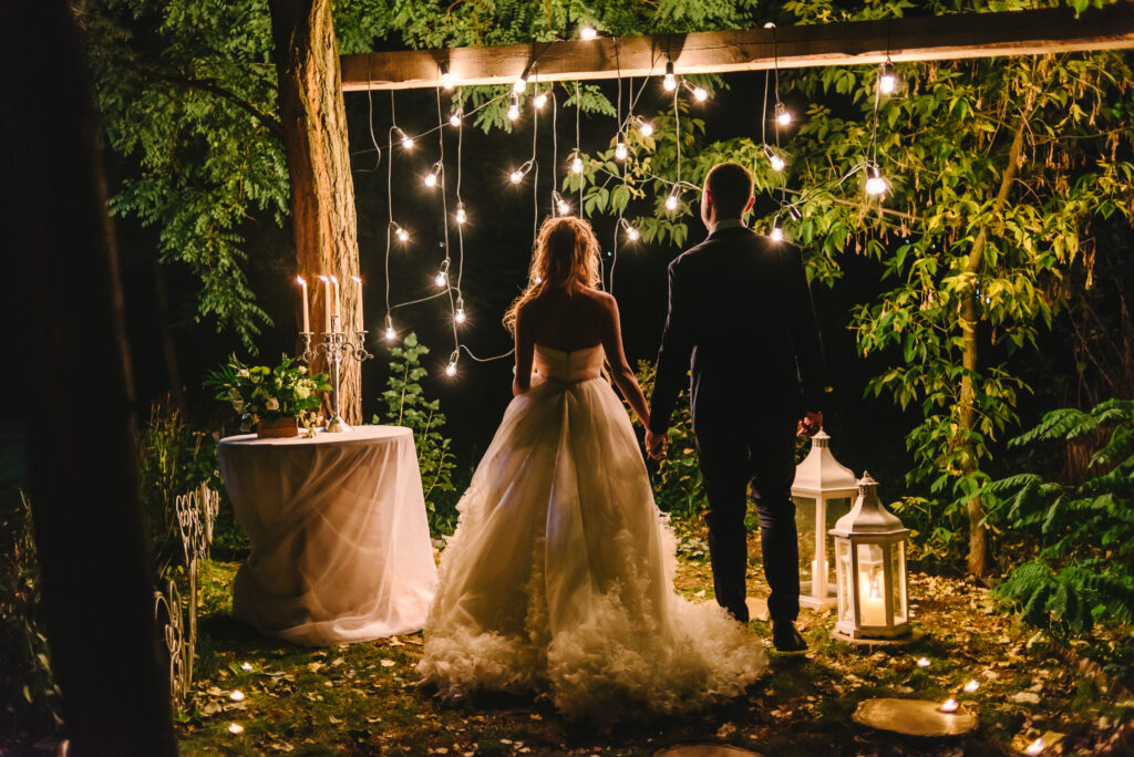 Night wedding ceremony with candles, lanterns and lamps on tree. Bride and groom holding hands on background of bulb lights, back view. Beautiful young couple standing under a tree at night