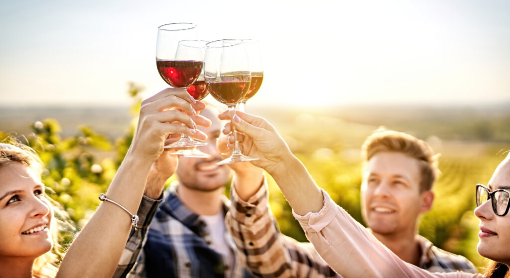 Happy people enjoying harvest time together at farmhouse winery countryside - Youth and friendship concept - Toasting red wine glass at vineyard before sunset - Focus on the wine glass